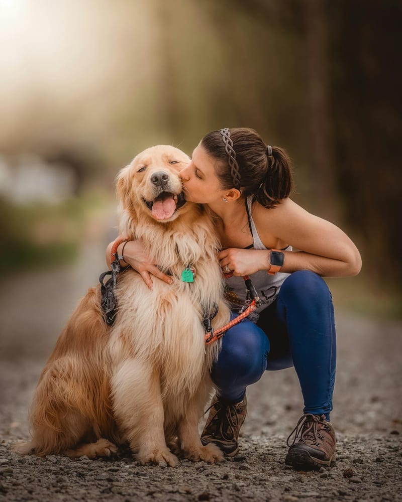 A woman crouching on a path to kiss a happy golden retriever